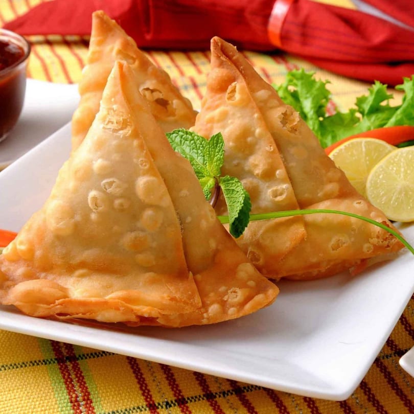 Golden-fried Annapoorani samosas on a white plate, garnished with mint, lemon, and lettuce.